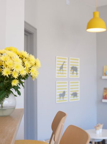 grey wall with four yellow pictures on it, yellow flowers and wooden chairs in the front and a yellow drop light