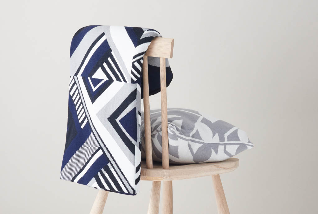 a wooden chair with a white, grey and blue pattern blanked over the backrest and a grey and white pillow on the seat