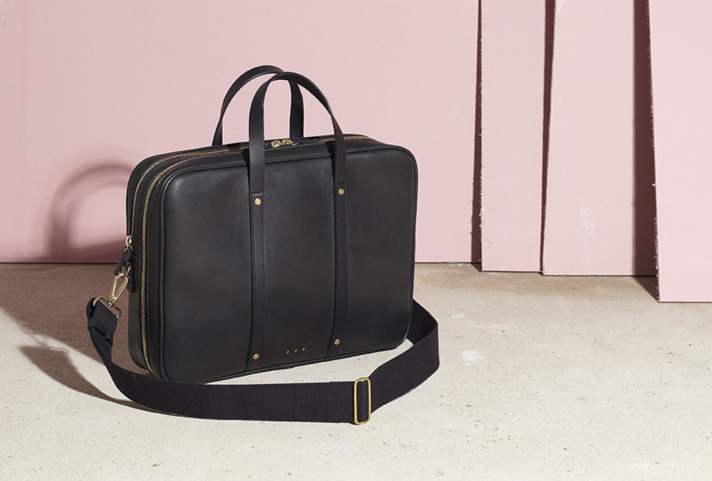 black men’s leather bag standing on a grey floor with pink walls